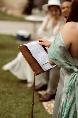Person in mint green dress holding papers at outdoor wedding with guests seated on grass.