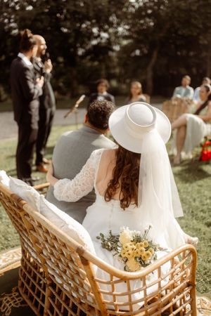 A couple sits together in a rattan loveseat during their outdoor wedding ceremony with guests in background.