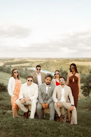 A stylishly dressed wedding party poses on a grassy hillside against a scenic countryside backdrop at sunset.