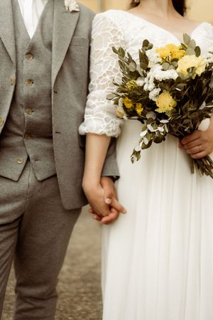 Wedding detail shot showing gray suit next to white lace dress with bouquet of yellow and white flowers.