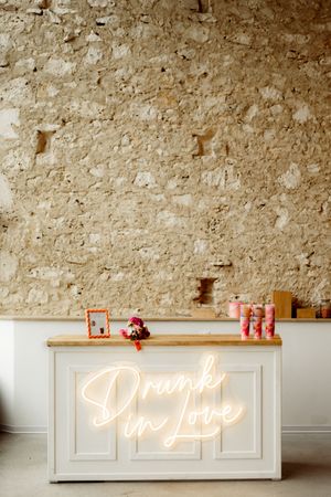 White bar counter with neon 'Drunk in Love' sign against a textured stone wall with decorative items on display.