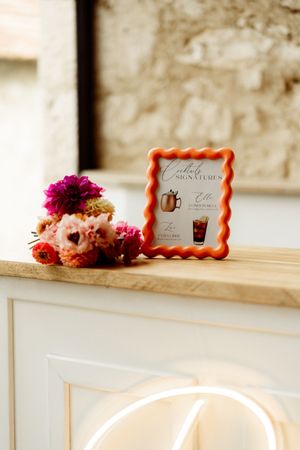 Rustic wooden shelf decorated with floral arrangements and ornate framed signs in coral tones against a neutral wall.