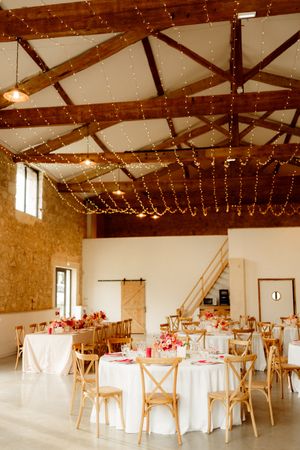 A rustic barn wedding reception venue with exposed wooden beams, string lights, and round tables with white linens.