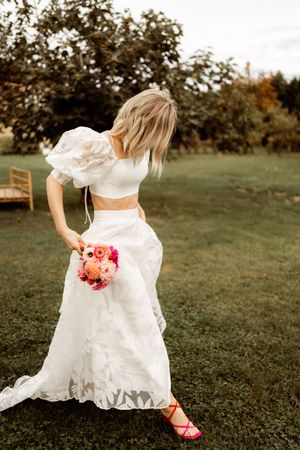 A person in a two-piece white wedding dress holds a vibrant pink and coral bouquet while standing on a grass lawn.