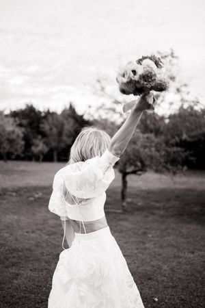 A figure in a white dress dancing joyfully in a garden field while raising their arms upward in celebration.