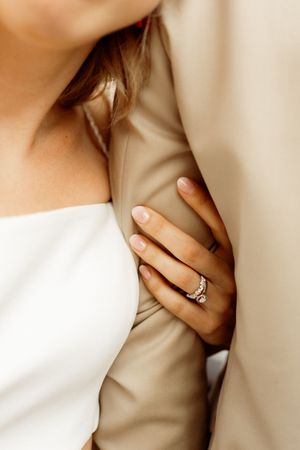 Close-up view of hands with diamond engagement ring against white dress and beige fabric background.