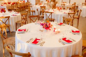 Elegant wedding reception tables with white linens, cross-back chairs, and red floral centerpieces set for a formal dinner.