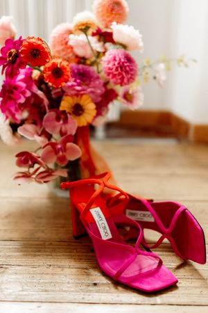 Red and pink heels beside a vibrant floral bouquet with dahlias and roses on a wooden table.