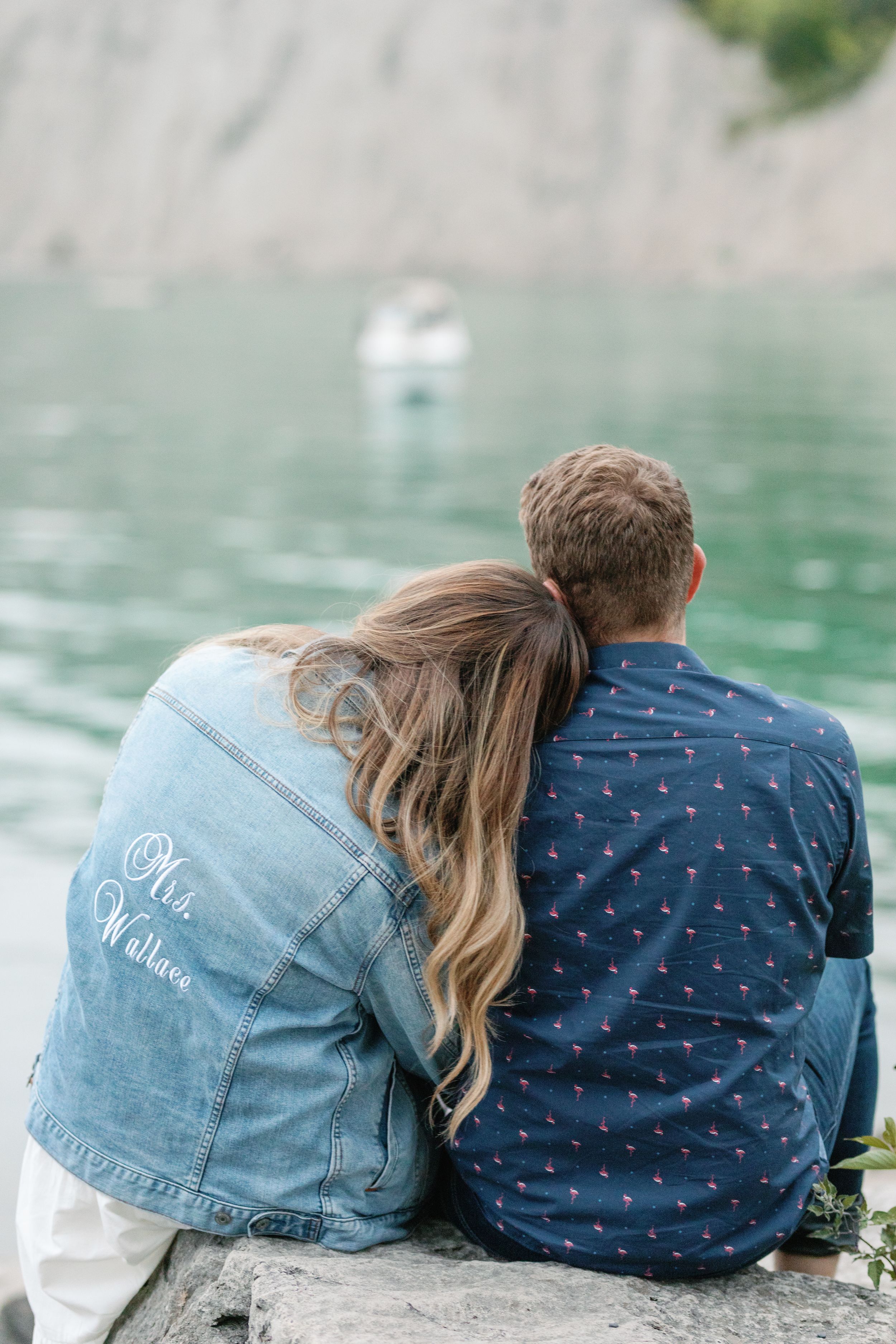 Couple wearing matching denim jackets sits by turquoise lake.