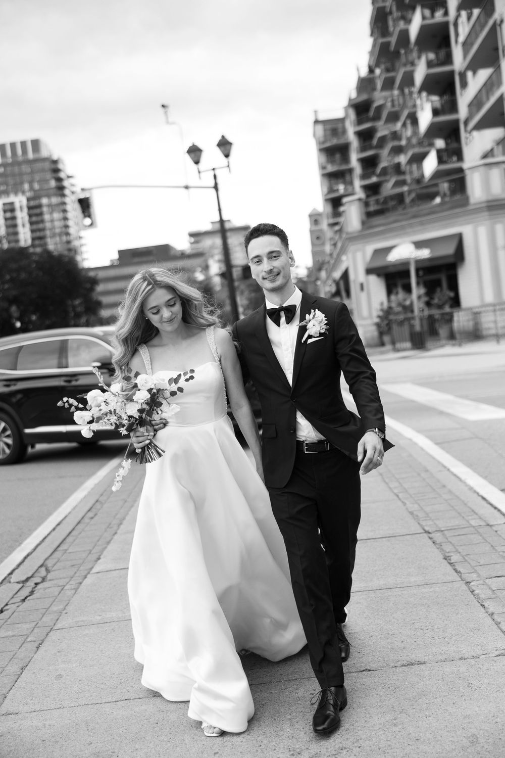 Black and white wedding photo series showing couple in formal attire posing playfully on city sidewalk.