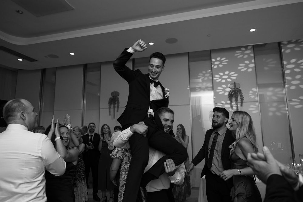 A person in a tuxedo is lifted up by wedding guests during a celebration in an elegant reception hall.