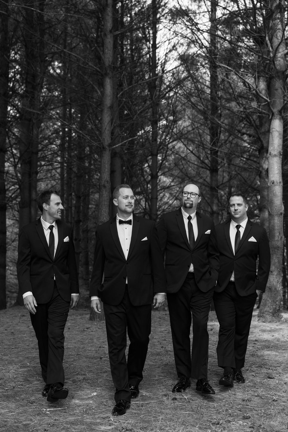 A black and white photograph of four men in formal suits walking together through a wooded forest setting.