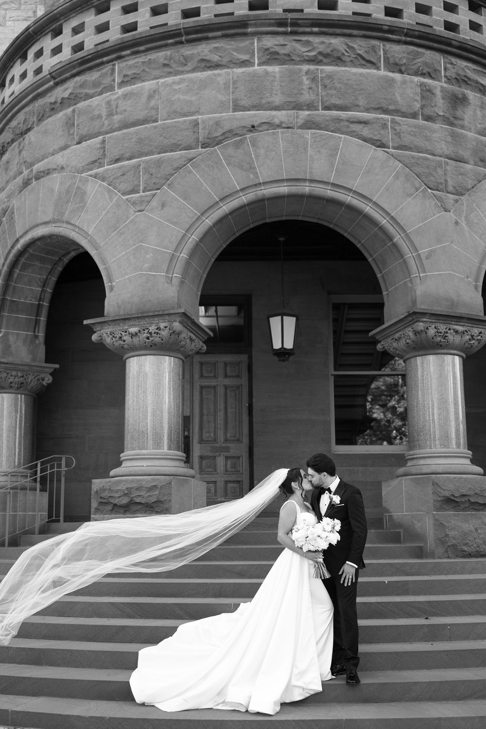 Romantic wedding couple embraces on stone steps of historic building with arched columns and flowing veil.