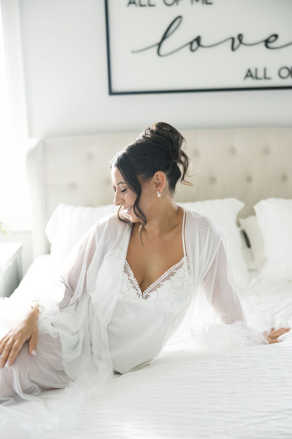 Bride in white lace lingerie and sheer robe lounges on white bedding with romantic decor in background.
