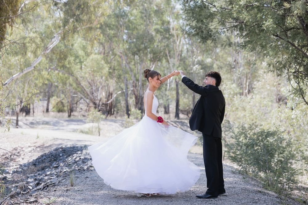 Groom twirling bride in white ball gown debutante outdoors among green trees on gravel path. location shepparton