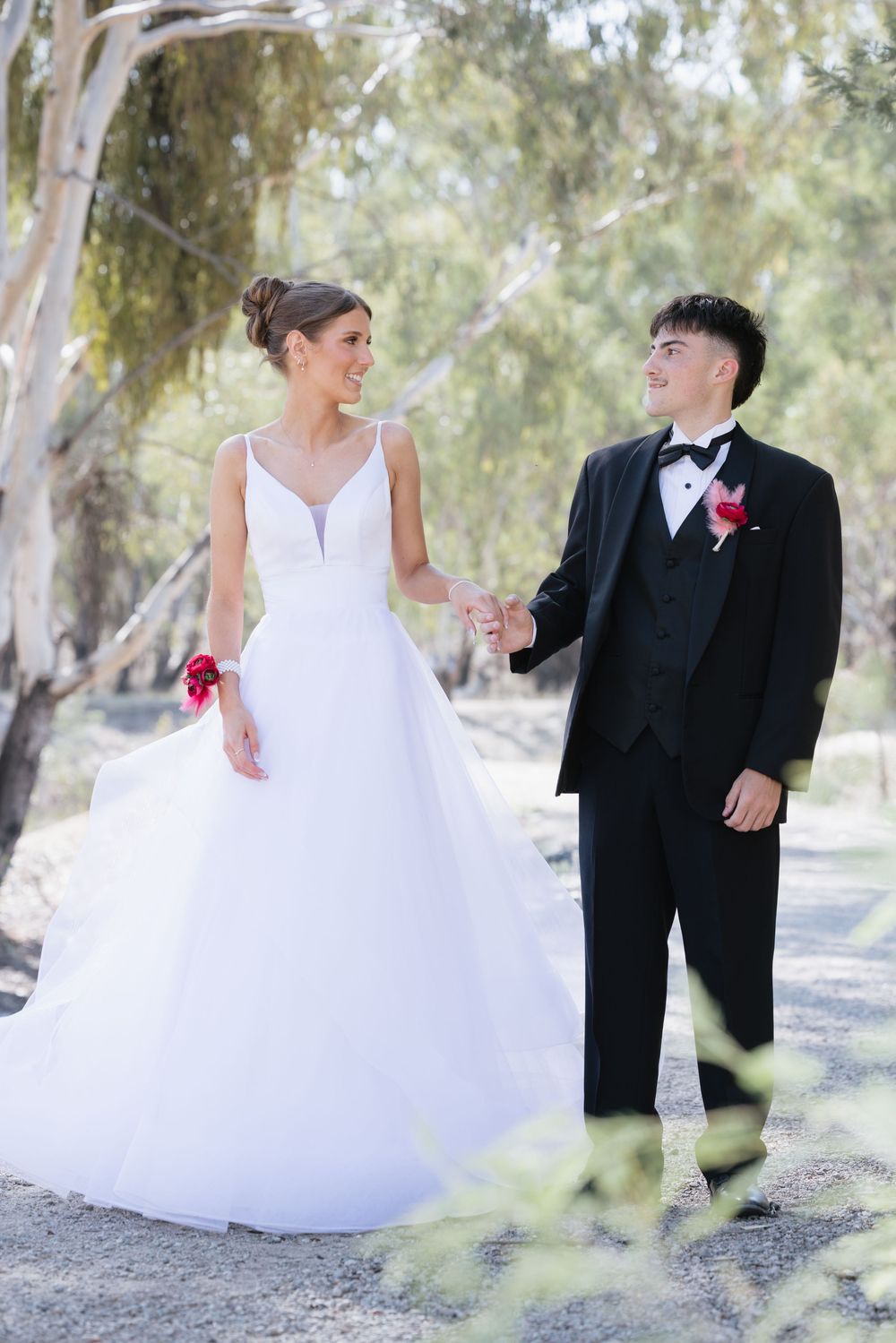 Girl in white gown and boy in black tuxedo holding hands outdoors among trees on their before debutante location Shepparton