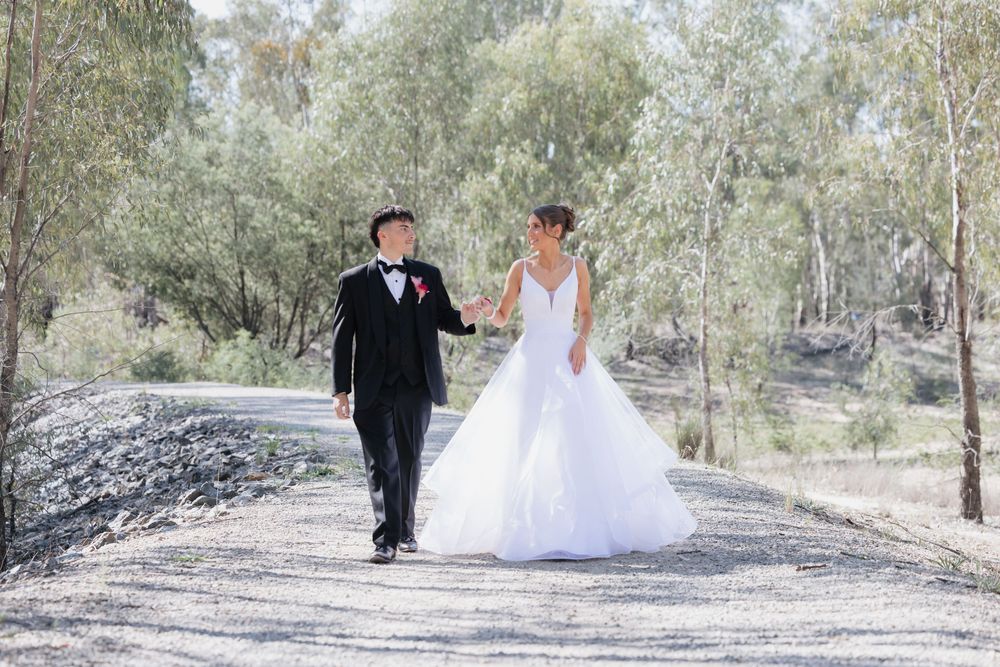 A deb couple walk hand in hand down a gravel path surrounded by trees on their wedding day.