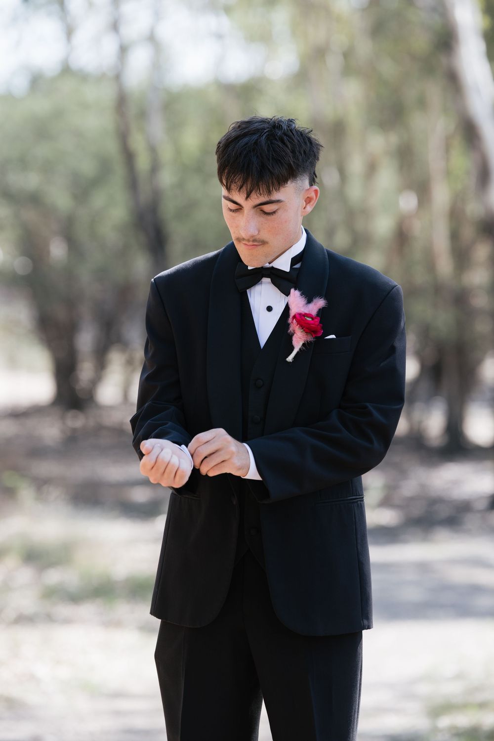 Young man in black tuxedo with red boutonniere adjusting cufflinks outdoors among trees.