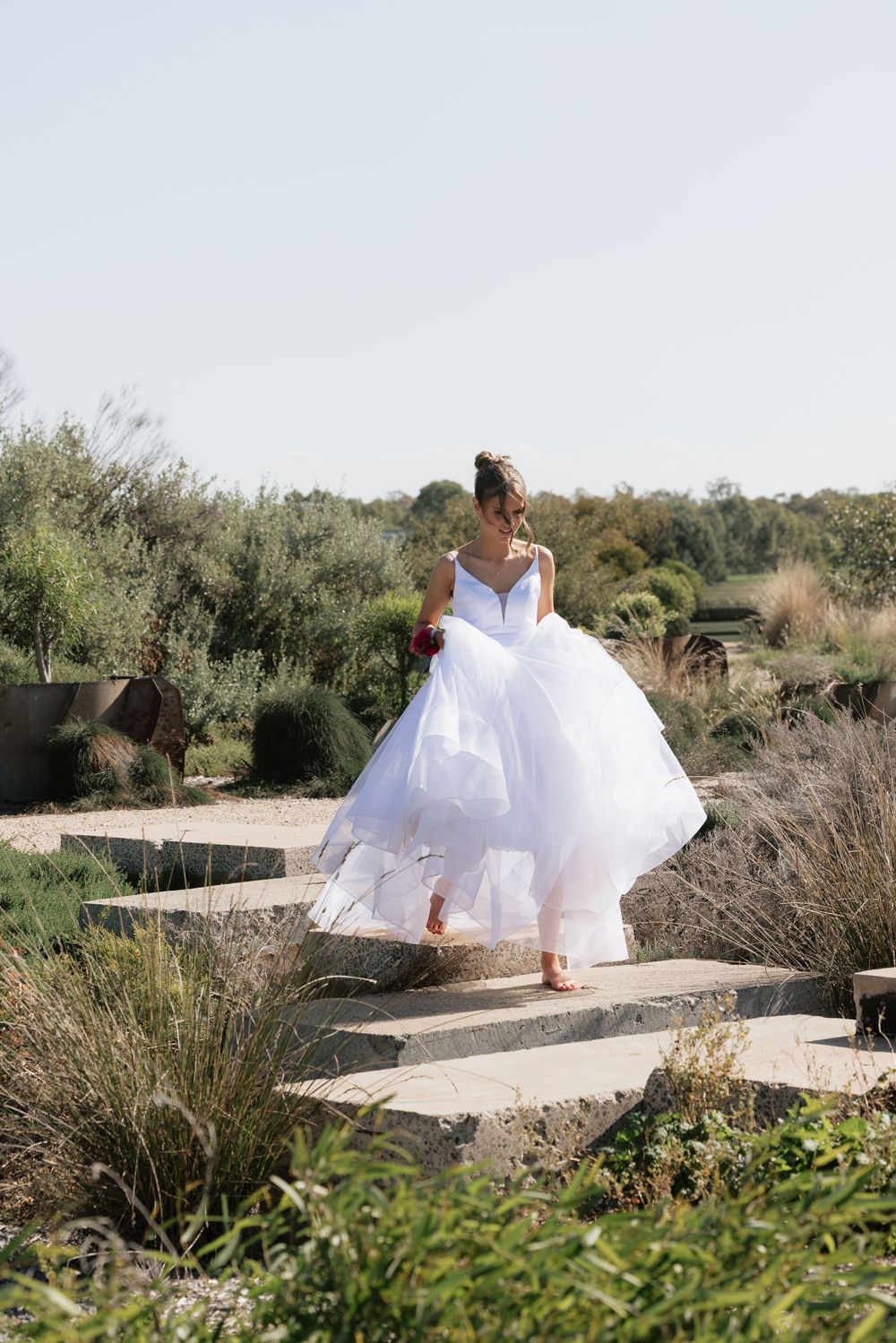 Girl in a white ballgown walks barefoot down stone steps in a lush garden setting, holding her dress and a bouquet. location shepparton