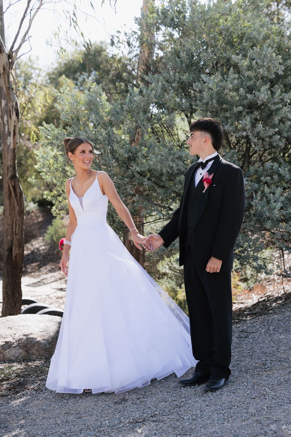 Young couple holding hands outdoors, girl in white debutante gown and guy in black tuxedo with red boutonniere. location shepparton