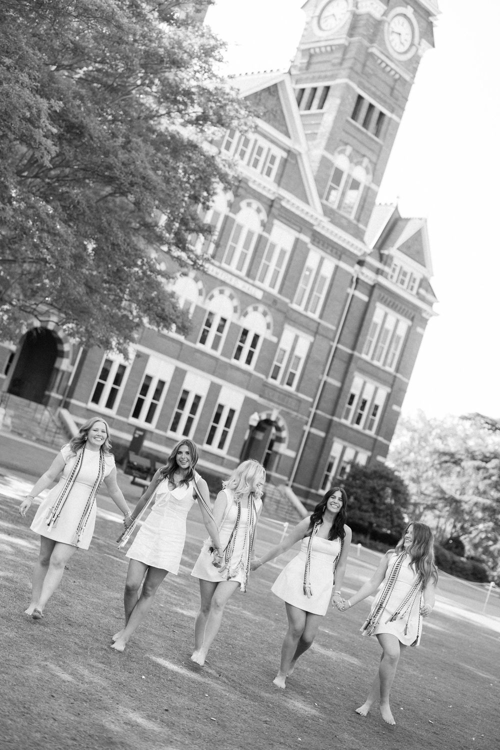A group of people walk in sequence across a lawn in front of a grand historic brick building with a clock tower.