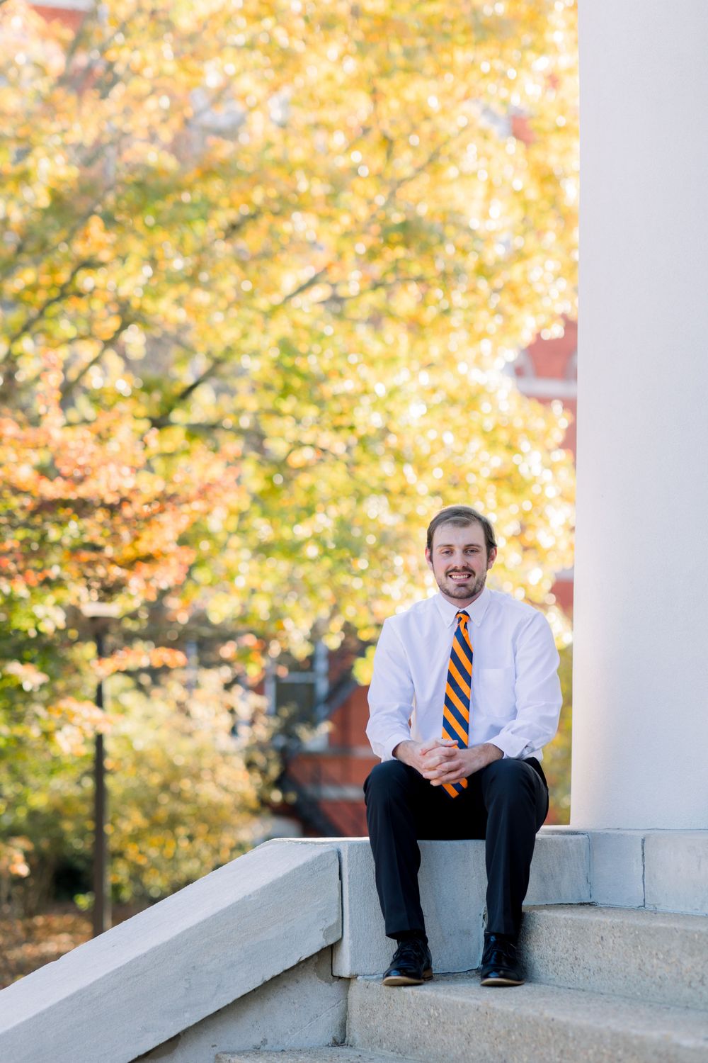 Graduate sits on concrete steps surrounded by autumn foliage and white columns.