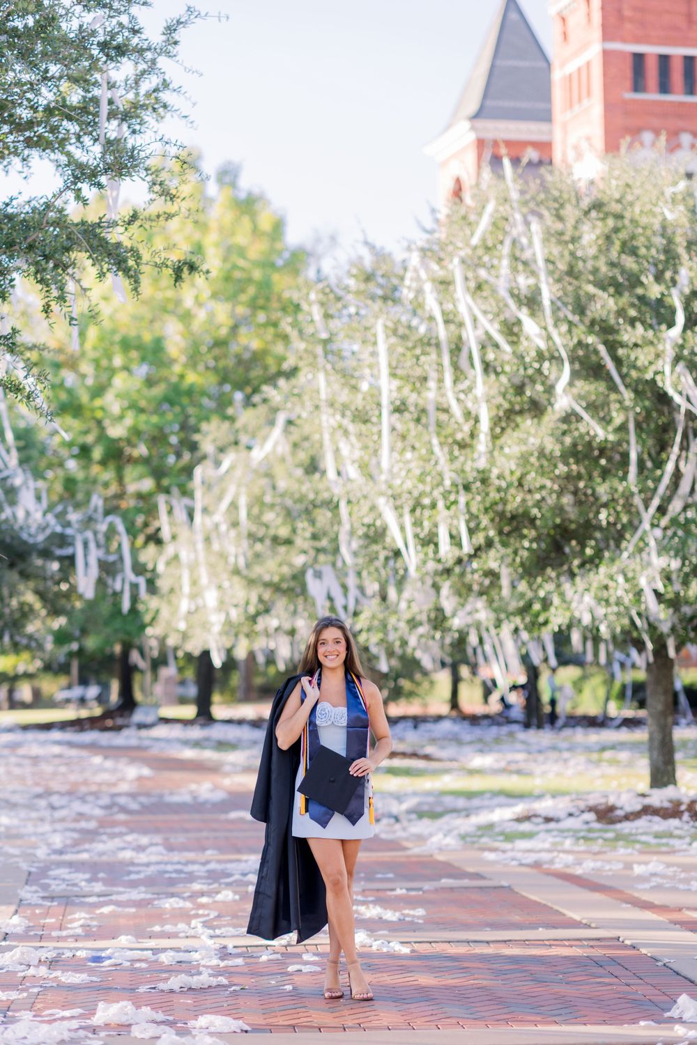 Graduate in white dress carries graduation gown while walking through campus.
