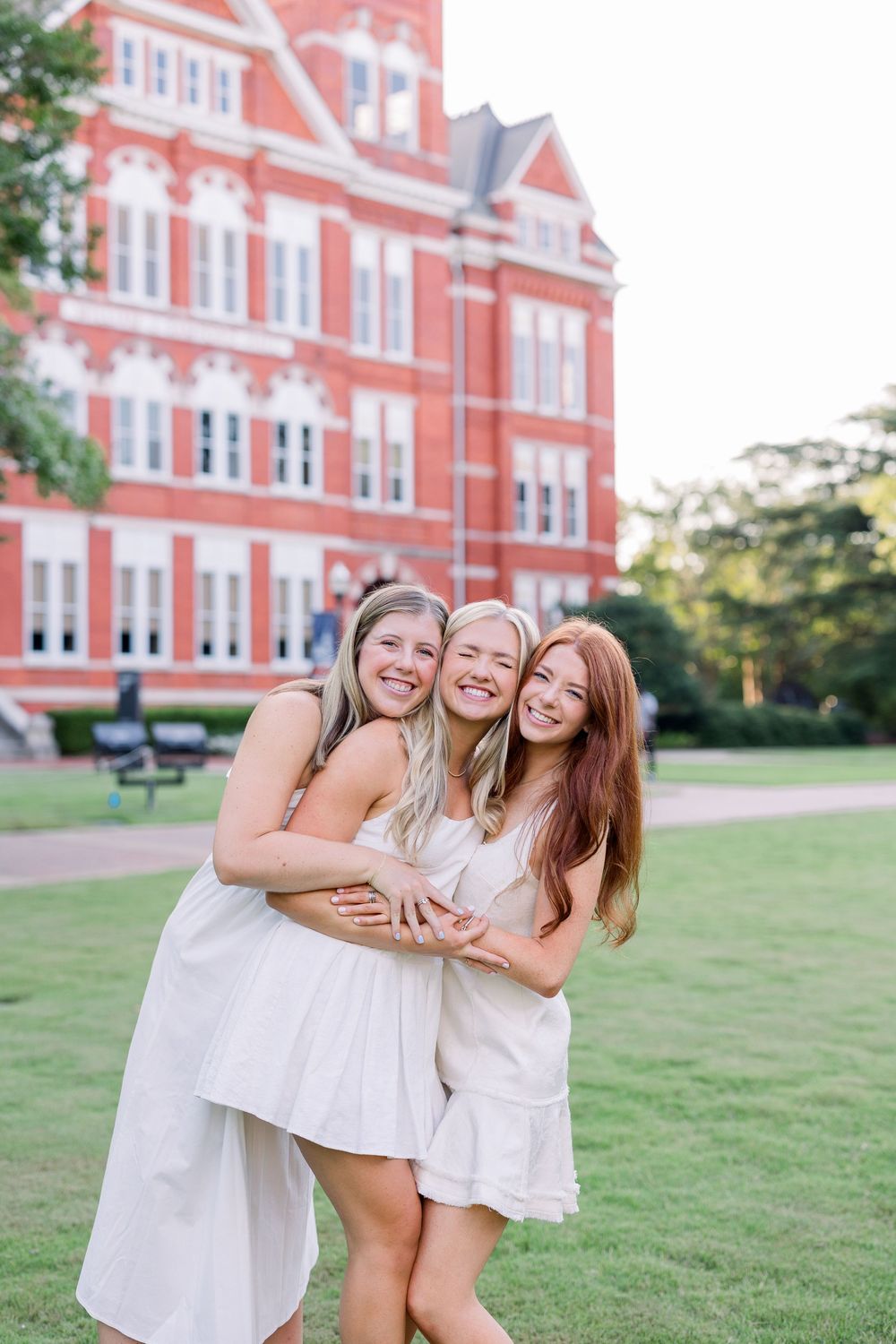 Three friends in white dresses laugh together on a grassy lawn in front of a historic red brick building.