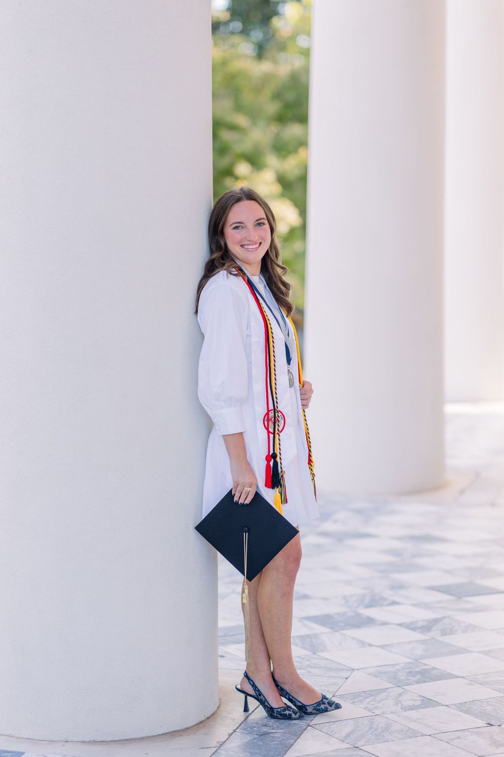 Graduate in white coat and colorful stole holds diploma while posing next to column during graduation photoshoot.