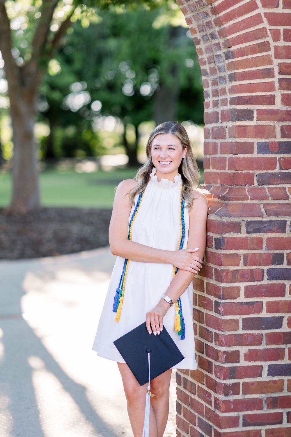 Graduate in white dress with honors cords leaning against brick wall.