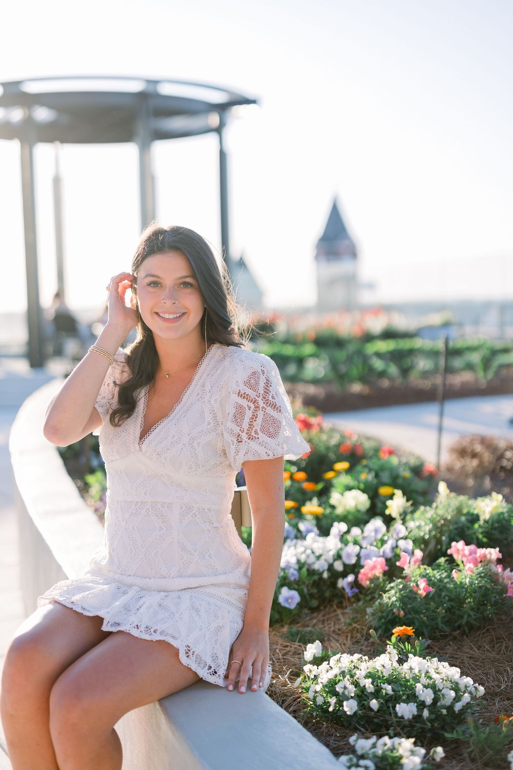 Woman in white lace dress sits on bench surrounded by colorful garden flowers with church spire in background.