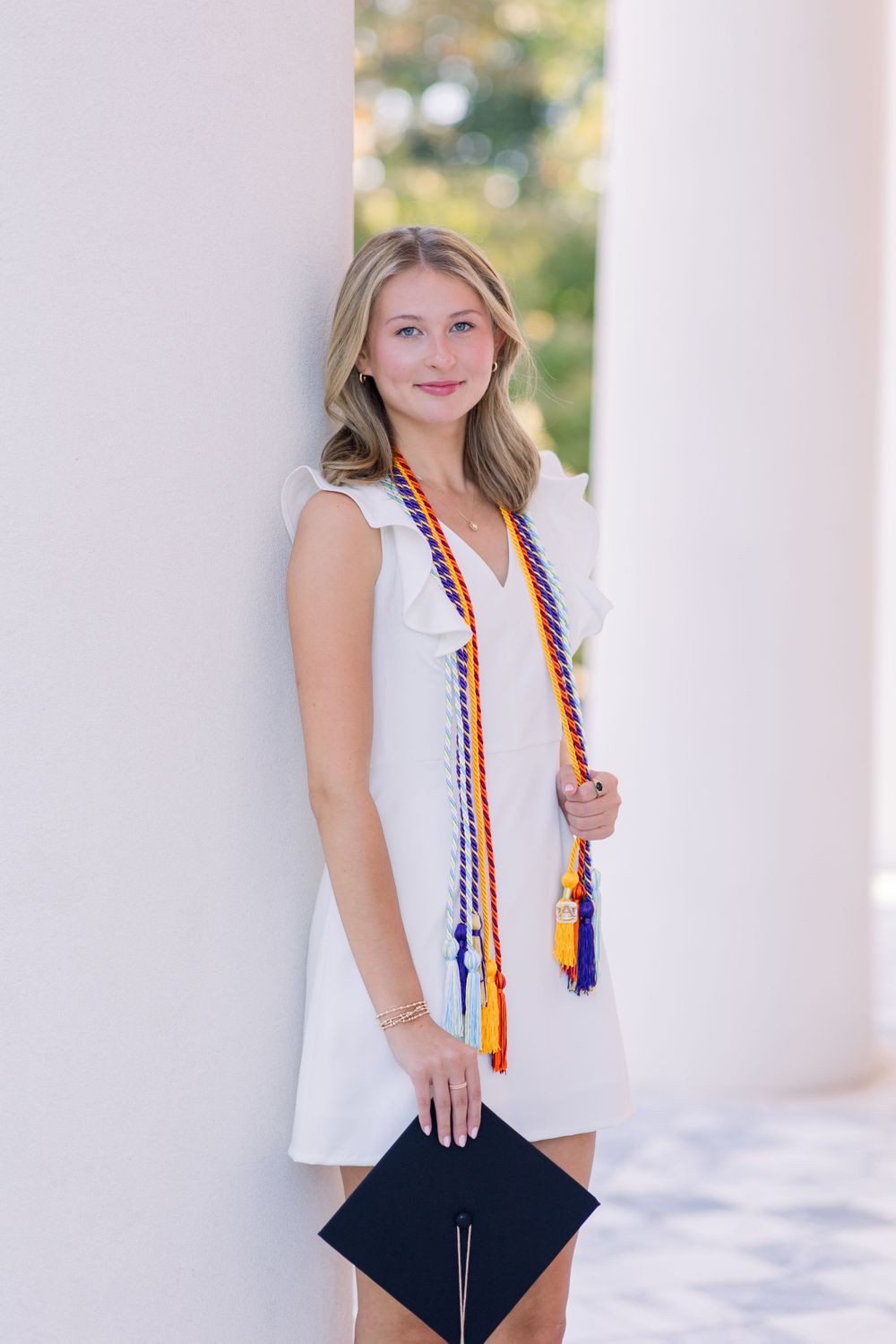 Graduate in white dress with graduation stole holds diploma against white column backdrop.