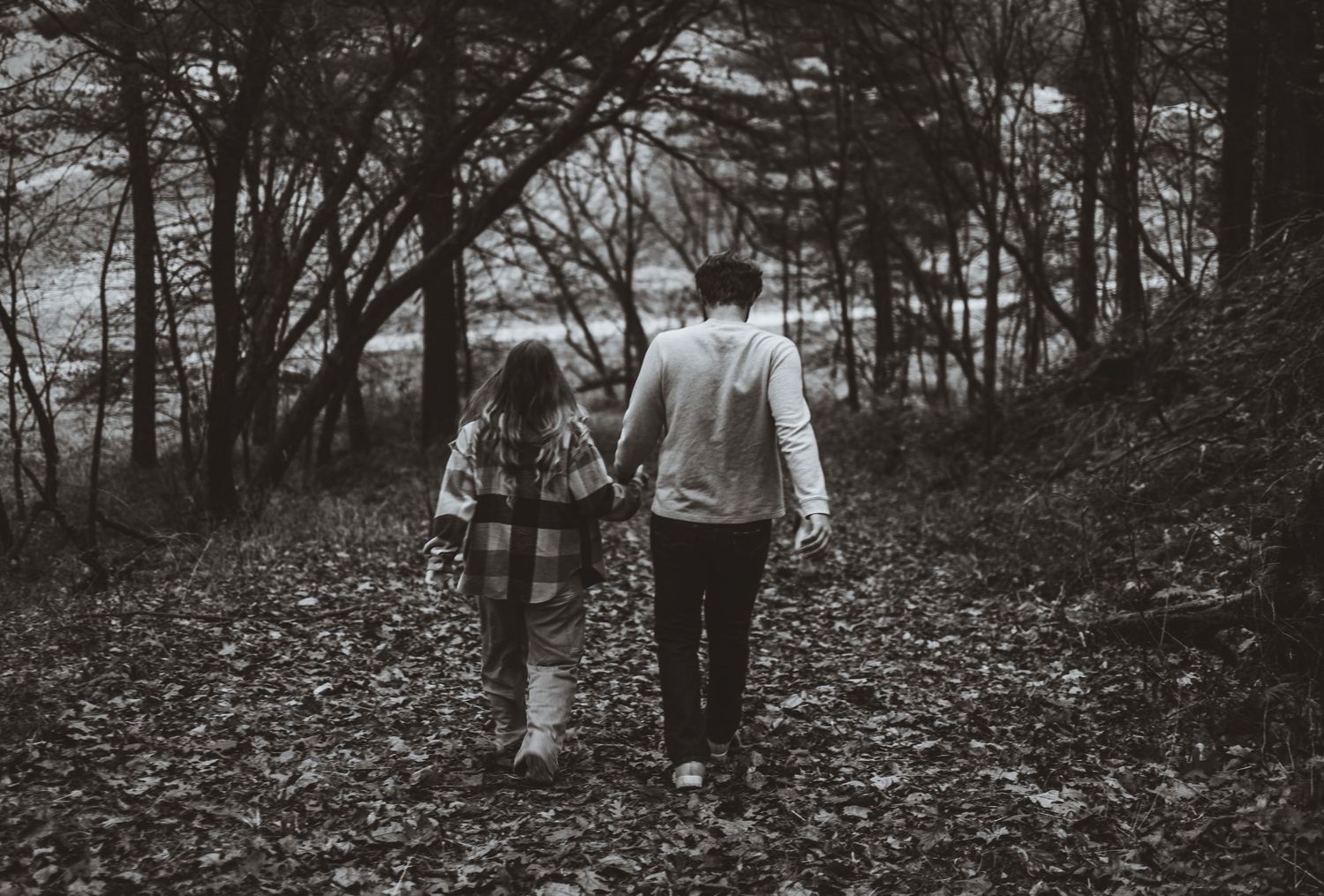 Black and white photo of a couple walking hand in hand through a wooded path covered in fallen leaves.