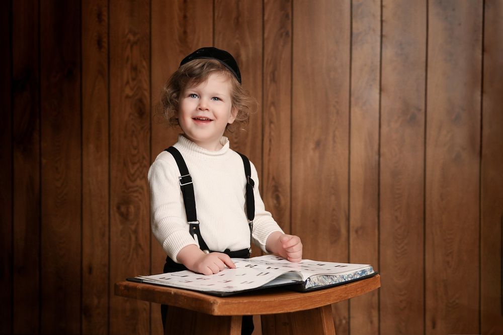 Young child wearing suspenders stands at wooden table looking at open book against wood paneled wall.