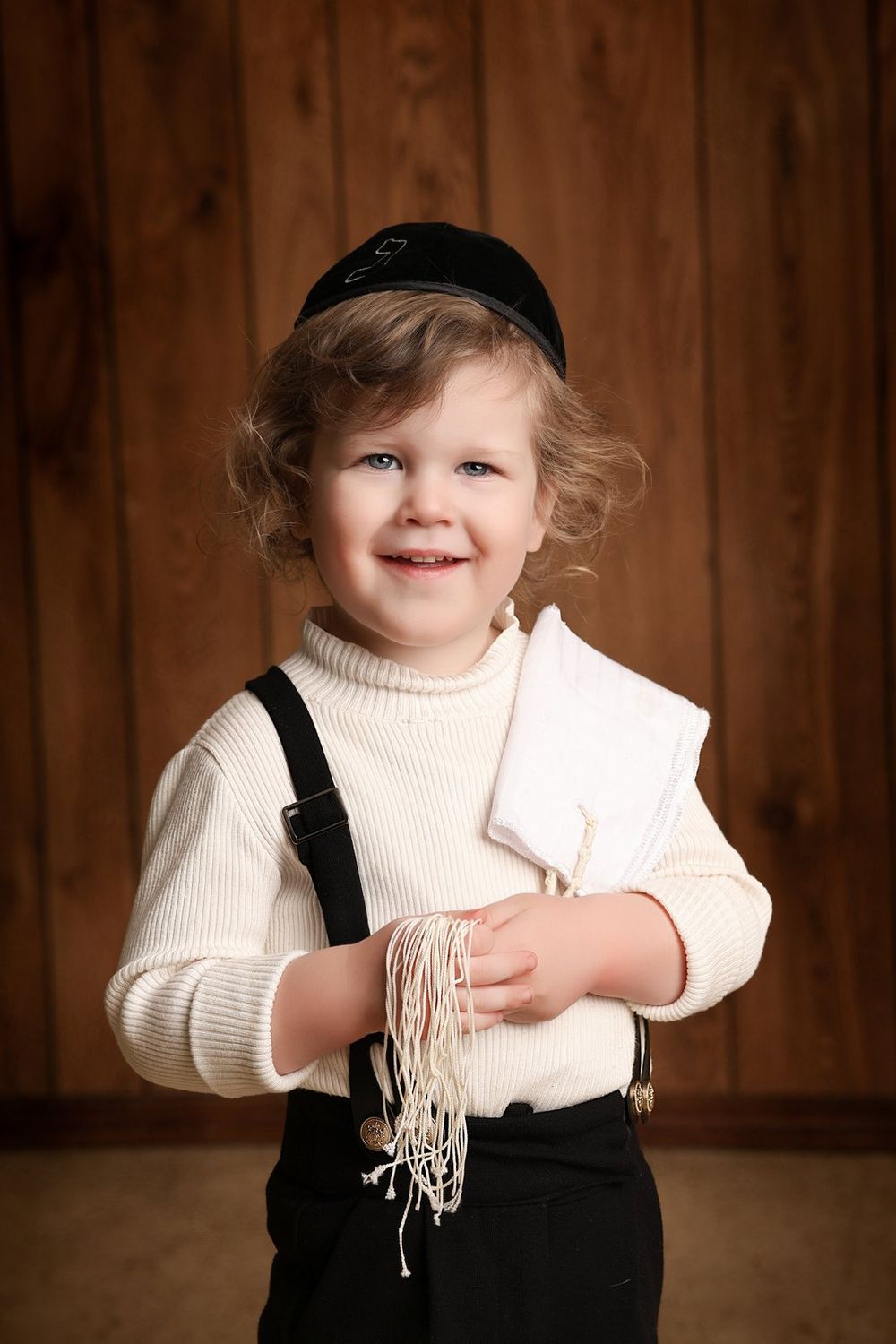 A toddler in a cream sweater and black suspenders smiles while holding fringe material against a wooden backdrop.