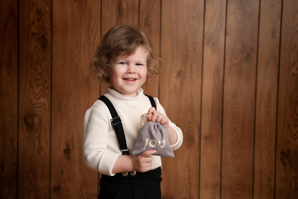 A young child wearing suspenders and a white sweater plays with a toy against wooden paneling.