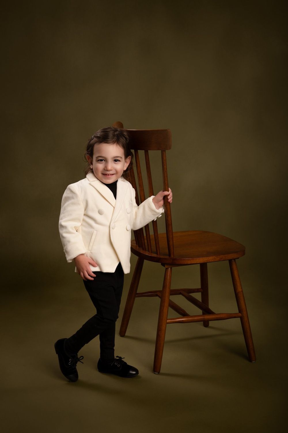 Small child wearing a white dinner jacket and black pants poses elegantly with a wooden chair against dark backdrop.