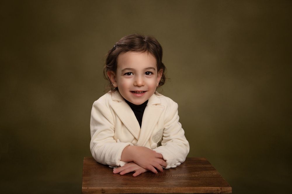 A child in a beige blazer and dark shirt poses at a wooden table against a dark backdrop.