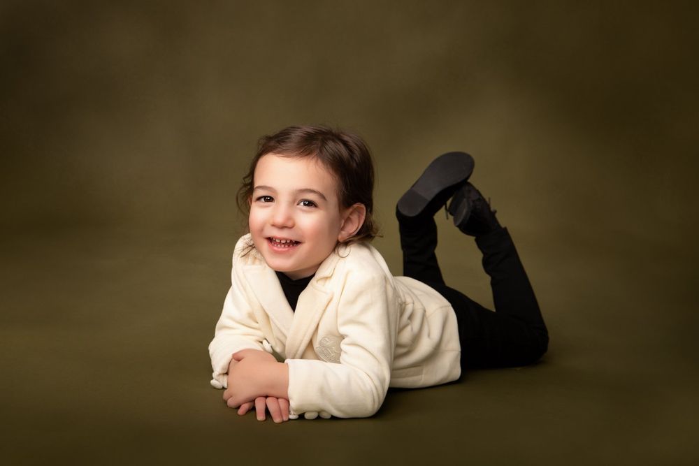 Child in a white sweater lying on dark background during professional studio portrait session.