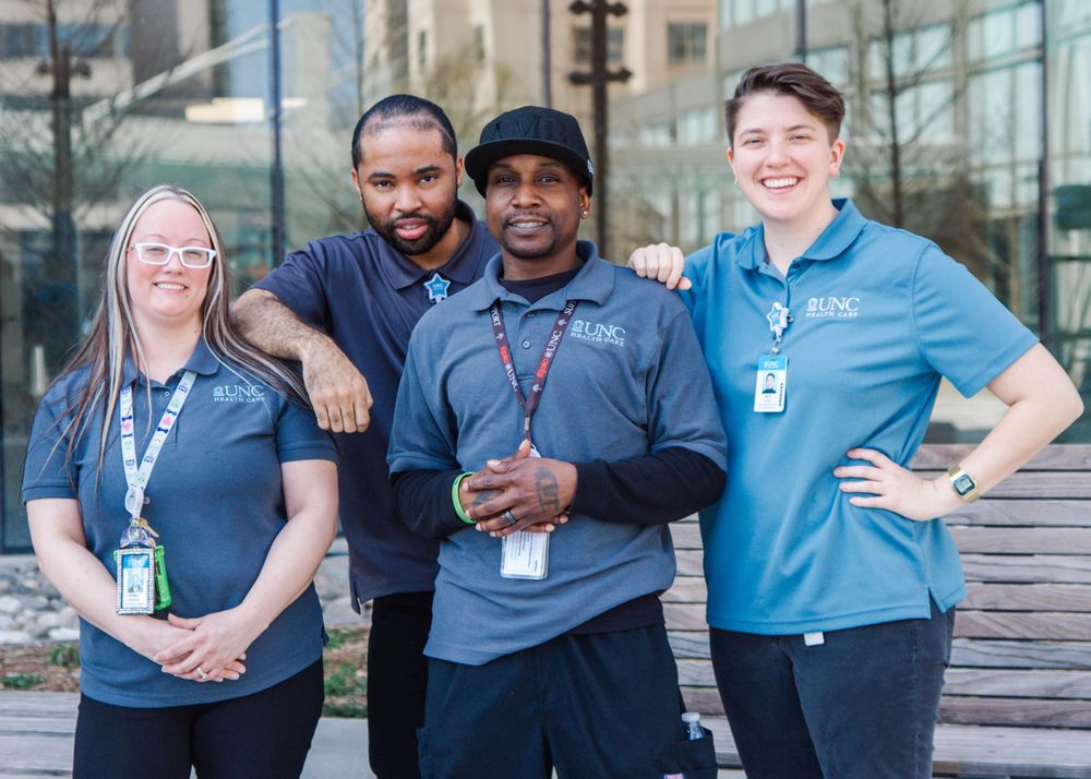 Four smiling healthcare workers in blue uniforms pose together outdoors near a modern building.