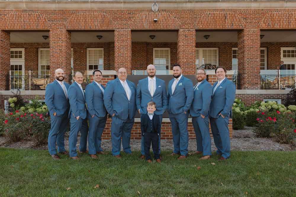 Wedding party wearing matching blue suits poses together in front of a brick building exterior.