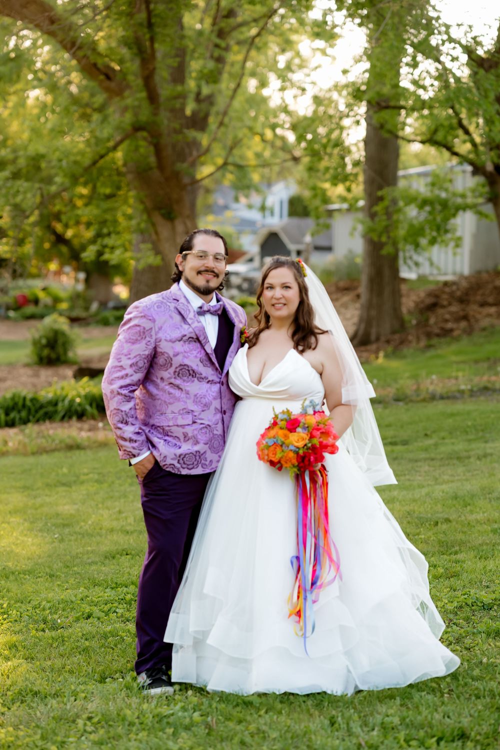 Bride in white gown holding colorful bouquet stands with groom in purple floral jacket on green lawn.
