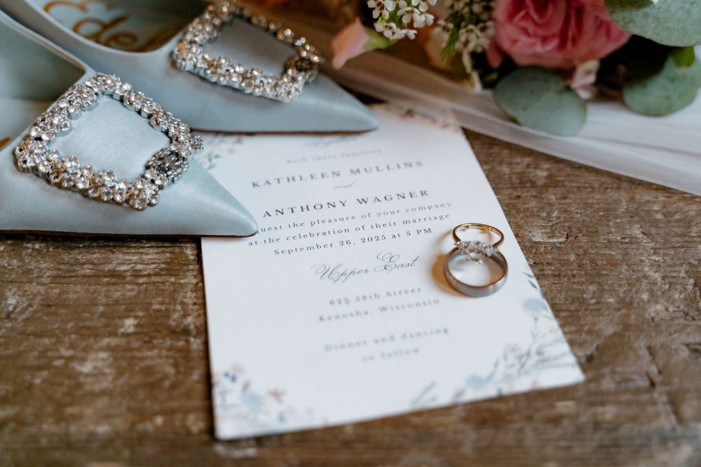 Wedding invitation with sparkly shoes and engagement ring displayed on rustic wooden surface with pink roses.
