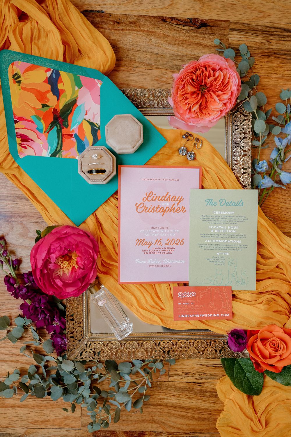 Colorful wedding invitation suite with pink and green cards, floral envelope liner, ring box, and fresh flowers on wood.
