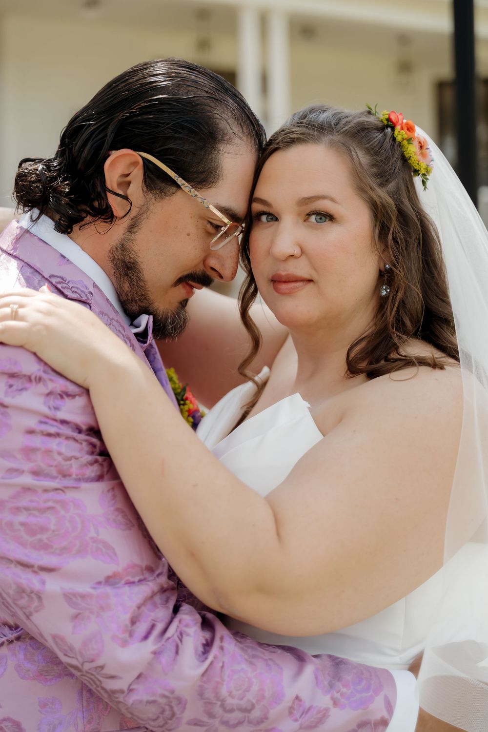 Bride and groom share an intimate forehead touch, she in a white gown with veil, he in a lavender floral suit.