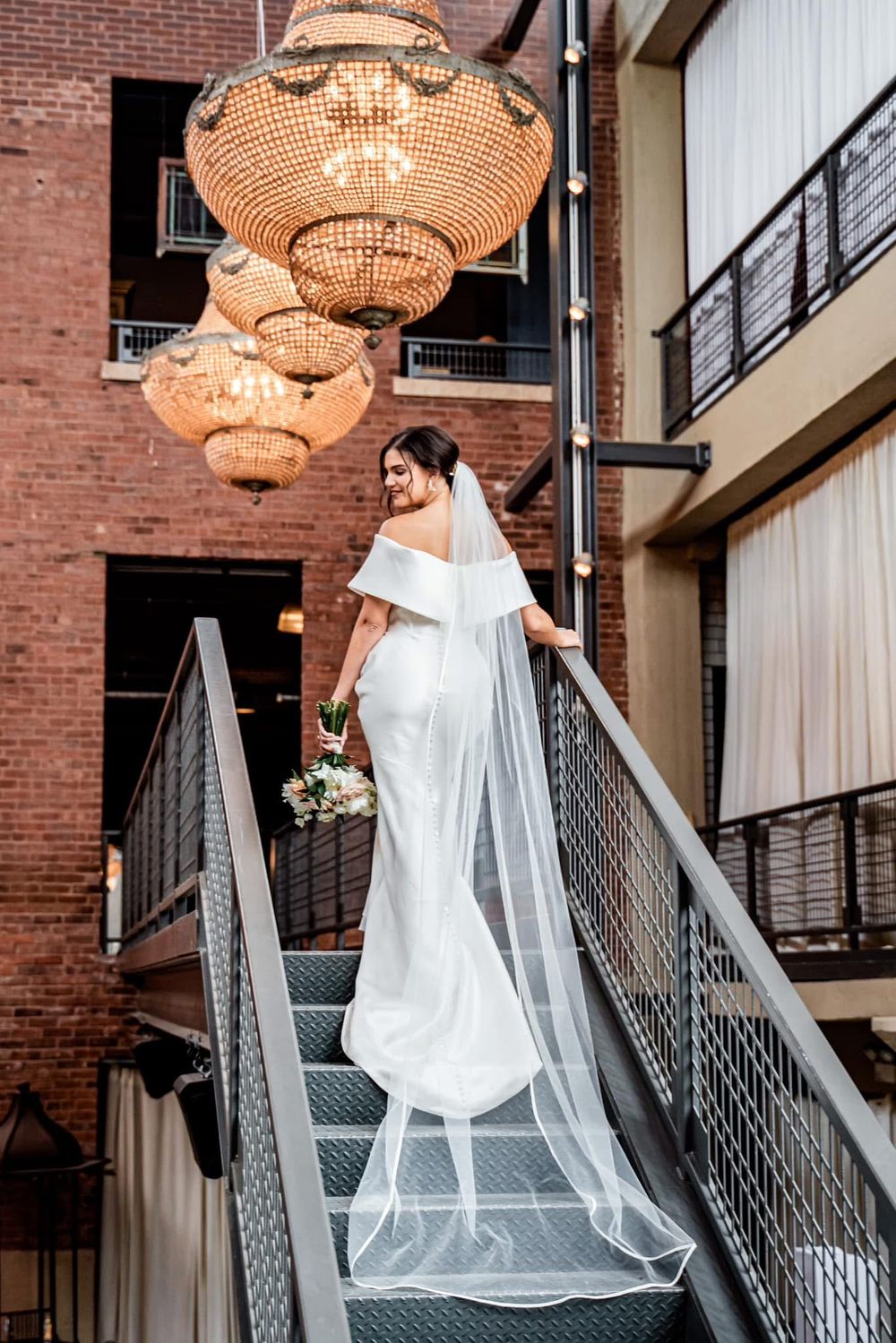 Beautiful bride in white gown descends industrial staircase under elegant crystal chandeliers holding bouquet.