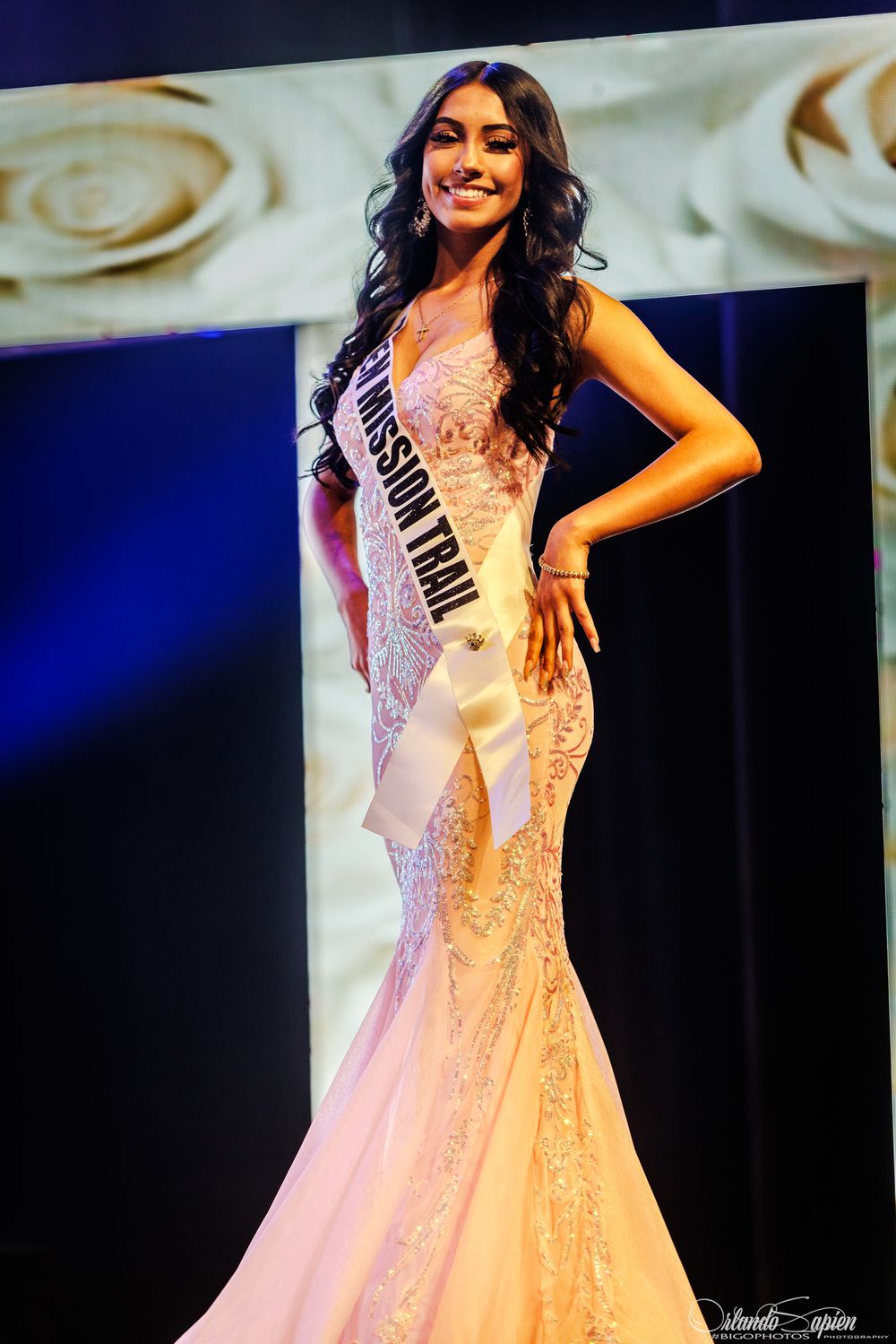 Elegant pageant contestants in white evening gowns on stage with blue backdrop.