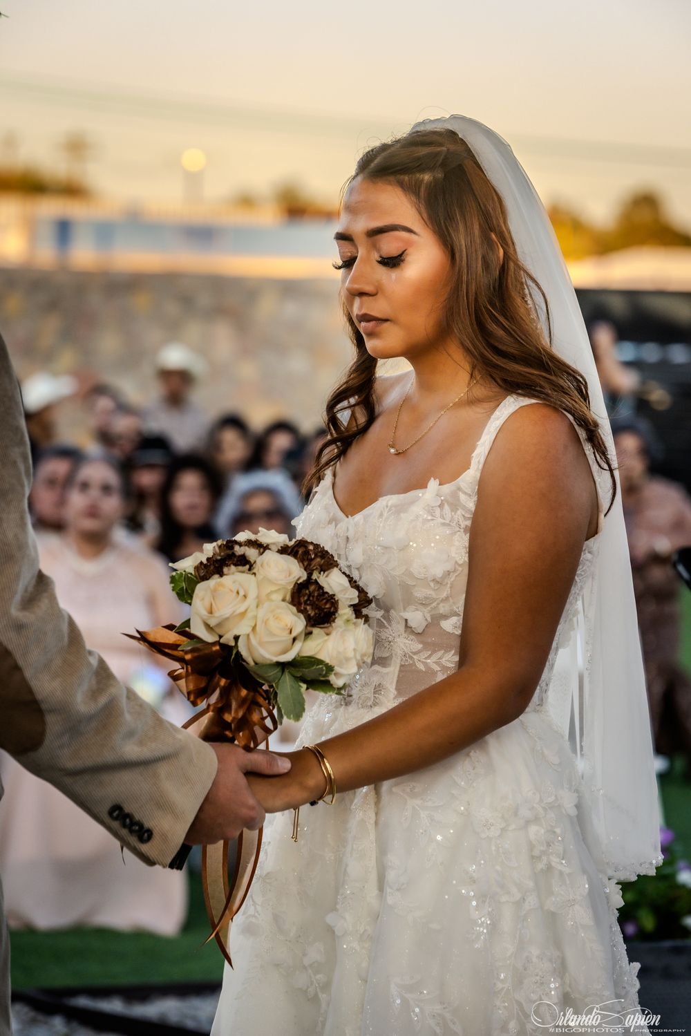 A bride in a white lace wedding dress holds a bouquet of white and burgundy flowers at sunset.