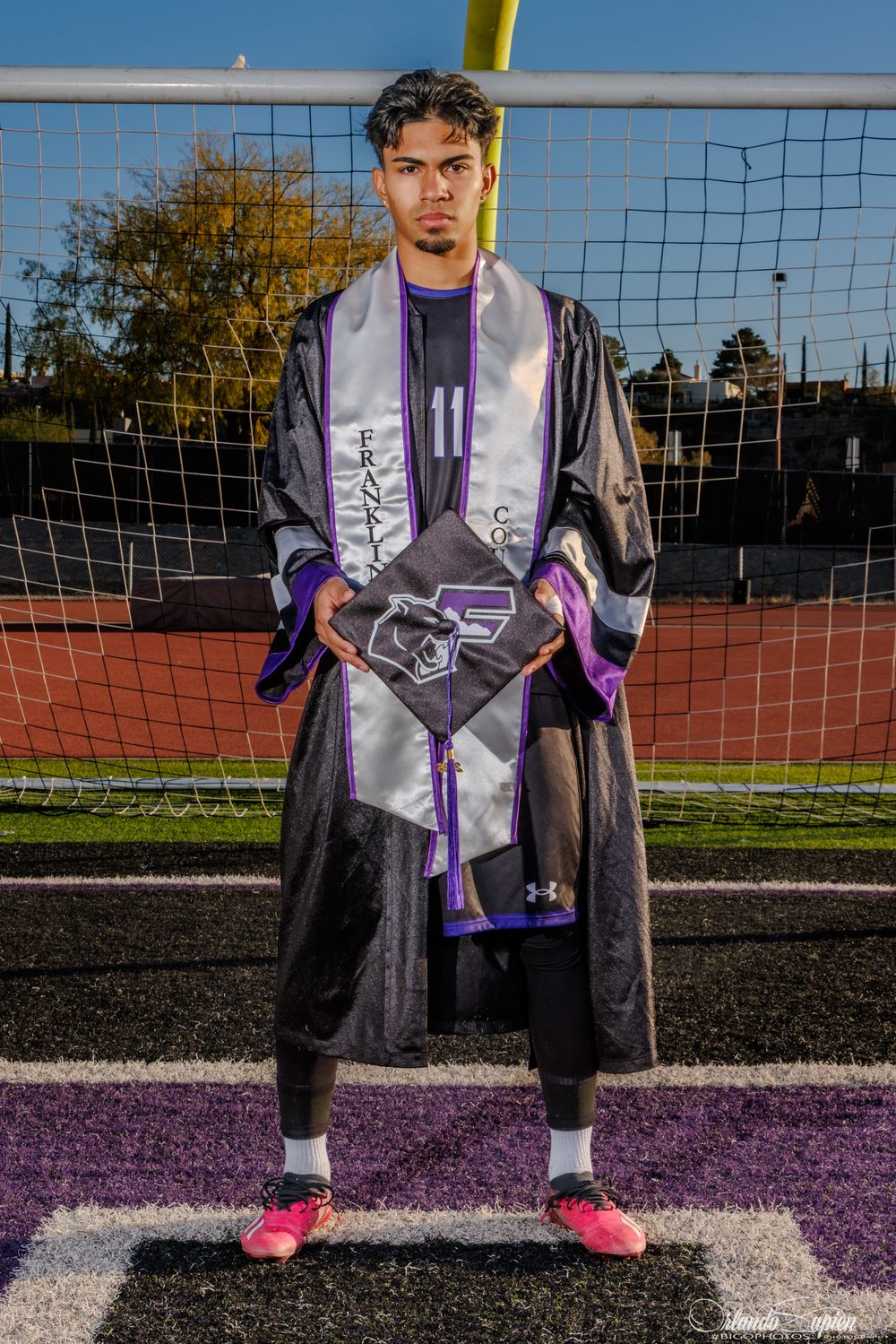 A soccer player wearing a purple and black uniform stands on a field with goal nets visible in the background.