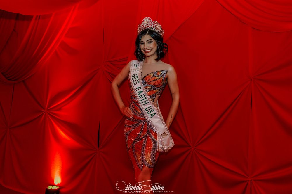 A pageant contestant in a red evening gown poses on stage with dramatic red lighting and backdrop.